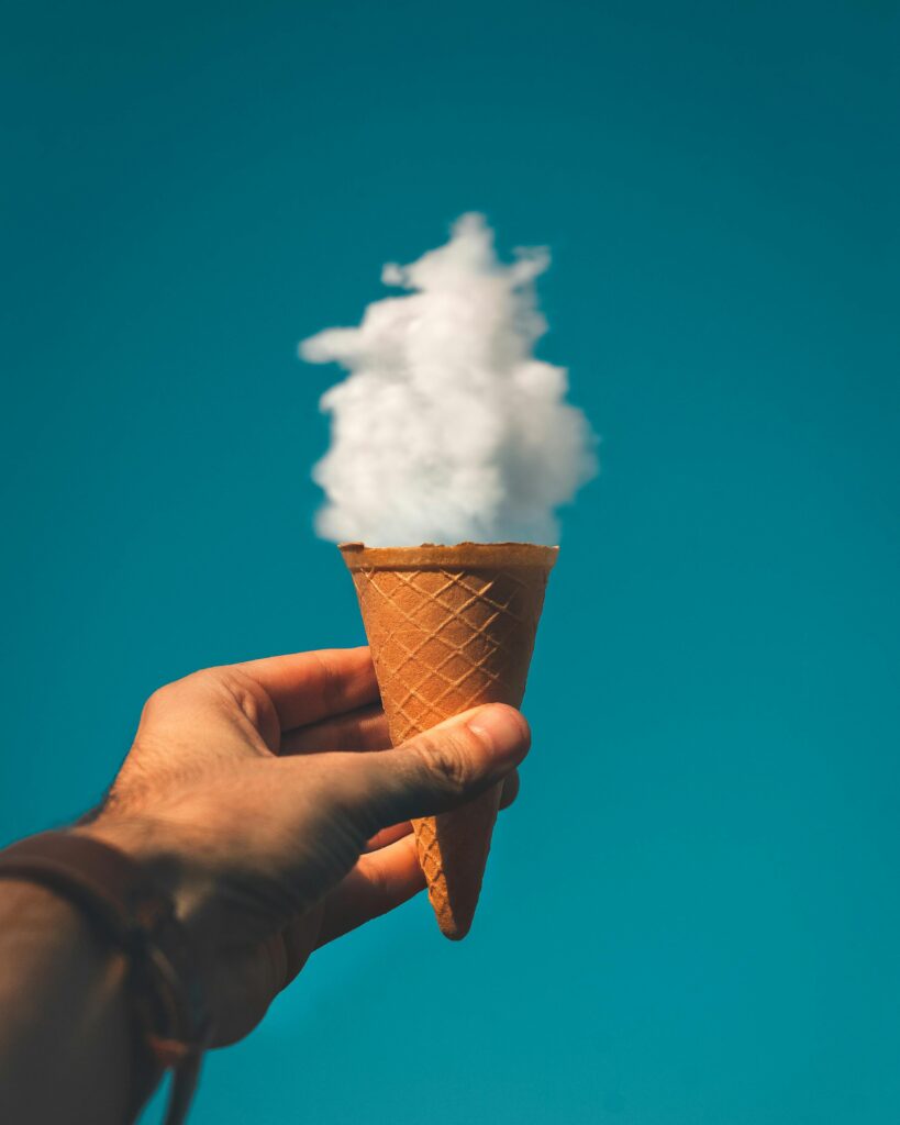 pexels-photo-1262302-1262302 Hand holding an ice cream cone with a cloud effect against a clear sky background.