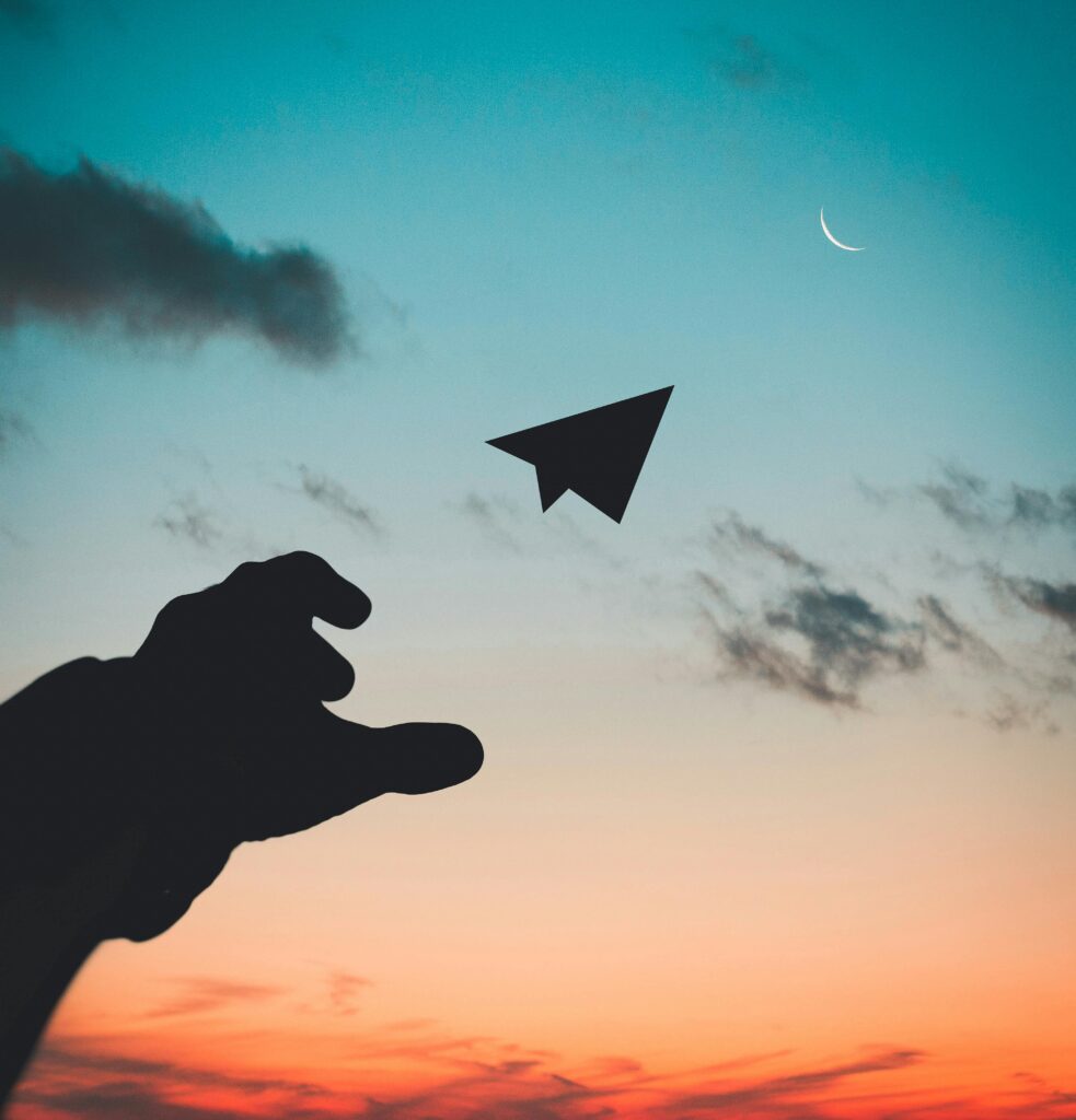 pexels-photo-1262304-1262304 A silhouette of a hand launching a paper plane against a colorful dusk sky with a crescent moon.